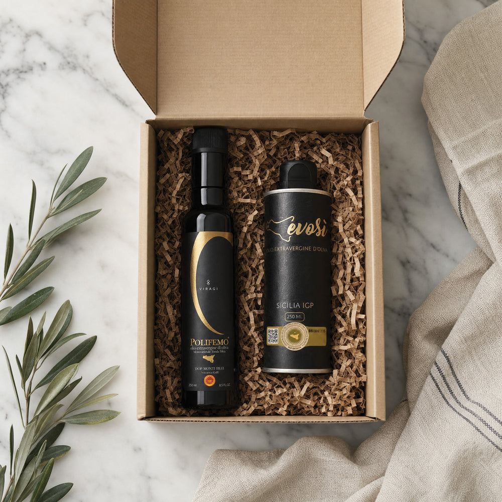 An open kraft gift box filled with brown crinkle paper packaging, containing two black bottles of extra virgin olive oil. The box is styled on a white marble surface with green olive branches on the left and a beige linen cloth on the right.