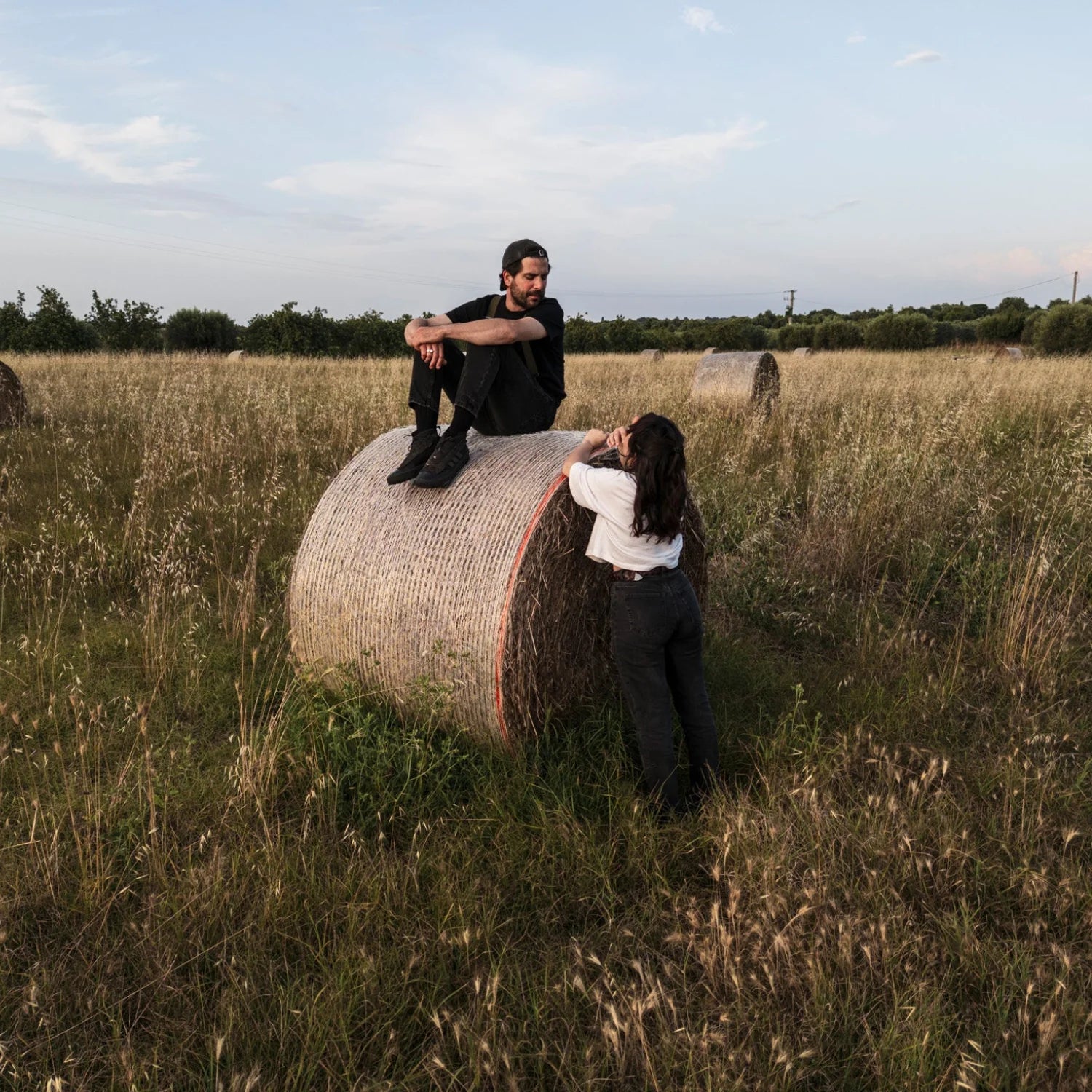Two people sitting on a hay bale in a field with a clear sky.